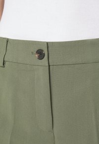 I pantaloni verde oliva presentano una parte anteriore piatta, una texture sottile, una chiusura a bottone visibile e una cucitura ordinata lungo la vita.