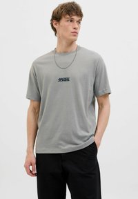 Gray short-sleeve t-shirt with a round neckline and a printed logo in black. The fabric appears smooth with a slight sheen. Paired with black pants.