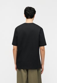 Άτομο με κοντά μαύρα μαλλιά που φοράει μία απλή μαύρη κοντομάνικη μπλούζα και χακί παντελόνι, γυρισμένο με την πλάτη σε λευκό φόντο.