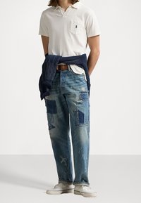 Maglietta polo bianca, jeans in denim chiaro strappati con vari patchwork e sneakers bianche, completando un look informale. Maglione blu navy legato in vita.