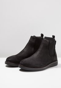 Une paire de bottes Chelsea en suède noir avec des panneaux latéraux élastiques, des tirettes et des semelles en caoutchouc basses, présentée sur une surface blanche.