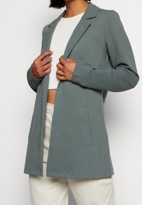 Blazer grigio chiaro a coste con taglio sartoriale, maniche lunghe e rever a incavo. Presenta una tasca frontale singola e design a fronte aperta.