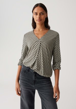 Vrouw in een zwart-wit geometrisch V-halsblouse met 3/4 mouwen en zwarte hoge taille jeans tegen een effen achtergrond.