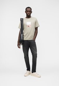 Λαδί πράσινη κοντομάνικη μπλούζα με τετράγωνο γραφικό σχέδιο, σκούρα τζιν και μπεζ αθλητικά παπούτσια. Το μοντέλο κρατάει μια μαύρη τσάντα ώμου, στέκεται μπροστά σε λευκό φόντο.