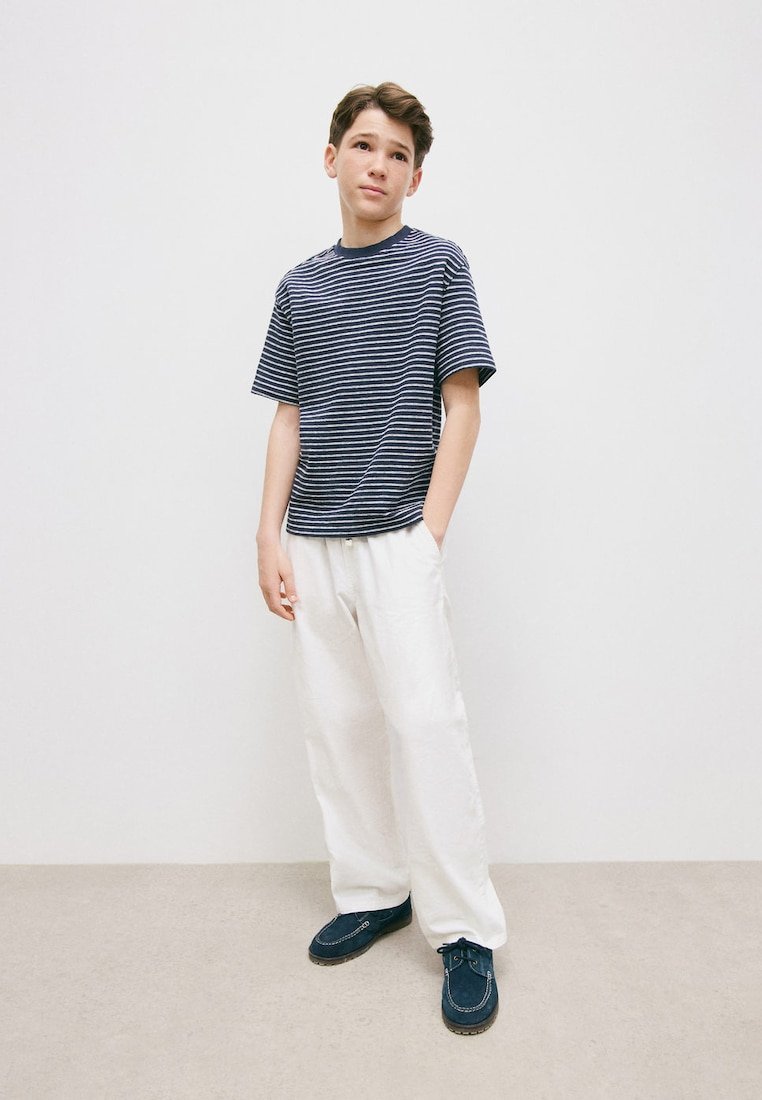 Garçon debout, les mains dans les poches, portant un t-shirt rayé bleu marine, un pantalon blanc ample et des chaussures décontractées bleu marine, devant un mur blanc.