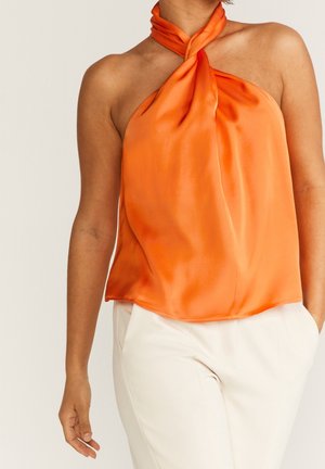 Kobieta w pomarańczowym satynowym topie bez rękawów z wiązaniem na szyi i białych spodniach, ręka w kieszeni.