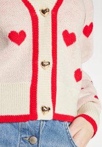 Cardigan lavorato a maglia color crema con accenti rossi, caratterizzato da motivi a cuore e bottoni lucidi a forma di cuore; abbinato a jeans di denim blu chiaro.