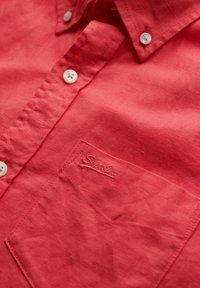 Camisa de botón color coral hecha de tela texturizada. Cuenta con un bolsillo con logotipo bordado y botones blancos. Diseño limpio y estructurado.