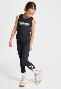 Svart ärmlös tröja med "hummel PERFORMANCE"-logotyp, kombinerad med svarta leggings med vita chevron-accenter på underbenet.
