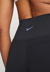 Leggings neri a vita alta in tessuto elasticizzato con cuciture visibili, caratterizzati da un piccolo logo Nike blu in vita. Texture liscia.