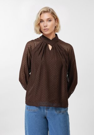 Ξανθιά γυναίκα που φορά διαφανές, σκούρο καφέ μπλούζα με υφή και ψηλό λαιμόκοψη, και ανοιχτό μπλε τζιν, στέκεται μπροστά σε λευκό φόντο.