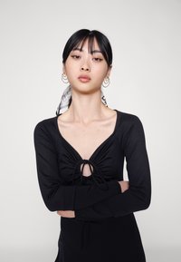 Top preto de mangas longas com decote em V e detalhe de cordão à frente. Modelo usa um lenço estampado no cabelo, brincos de argola e maquilhagem neutra.