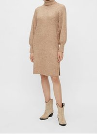Pletené šaty v béžové barvě s vysokým límcem, ribovanou strukturou, bočním rozparkem a knoflíkovými detaily podél levé strany. Stylizováno s světlými hnědými kotníkovými botami.