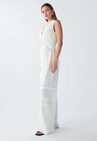 Mono blanco sin mangas con detalle de botones, cintura ceñida con cinturón y franjas horizontales transparentes en el dobladillo. El tejido parece ligero y transpirable.