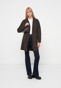 Cardigan marron à manches longues avec une ouverture sur le devant, texture semblable à de la laine. Associé à un jean évasé bleu foncé et des bottines noires.