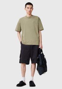T-shirt in cotone verde oliva con collo rotondo, abbinato a pantaloni cargo neri, calzini bianchi e scarpe slip-on nere, mentre tiene uno zaino blu navy.