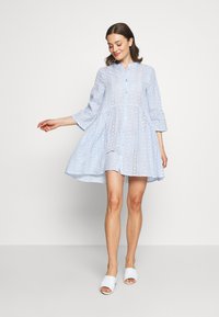 Vestido azul claro, com botões, mangas três quartos e recortes em forma de olhal, apresentando uma bainha rodada. Usado com sandálias brancas de slide.