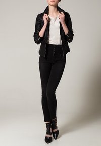 Svart läderjacka med texturerad yta, vit T-shirt, svart högmidjade jeans och svarta klackskor med remmar. Stående position.