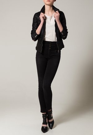 Czarna skórzana kurtka o fakturowanym wykończeniu, biała koszulka, czarne jeansy z wysokim stanem oraz czarne sandały na paski na obcasie. Pozycja stojąca.