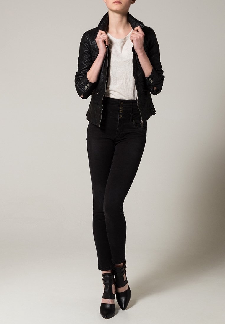 Svart läderjacka med texturerad yta, vit T-shirt, svart högmidjade jeans och svarta klackskor med remmar. Stående position.