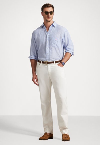 Gaiši zila un balta svītrainā krekla, balto biksu, brūna ādas josta, brūnās mokasīnu kurpes, melni saules brilles, stāvošā pozā.