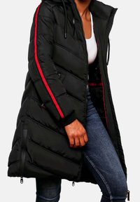 Femme portant une longue doudoune noire avec des bandes rouges sur les manches, partiellement zippée sur un haut blanc et un jean bleu.