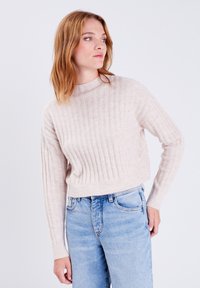 Beige gerippter Strickpullover mit hohem Kragen und langen Ärmeln, kombiniert mit hellblauen Jeans. Weicher Stoff und cropped Länge.