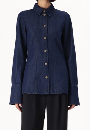 Vrouw met een donkerblauw denim overhemd met knopen, lange uitlopende manchetten en een puntige kraag, gecombineerd met losse zwarte broek.