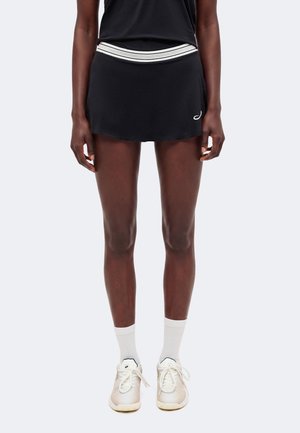 Mujer que lleva una falda deportiva negra con una cinturilla blanca a rayas, calcetines blancos hasta la altura del tobillo y zapatillas blancas con cordones, posando frente a un fondo liso.