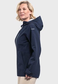 Marineblaue wasserdichte Jacke mit Kapuze, Reißverschluss vorne, elastischen Bündchen und Seitentaschen. Verfügt über weiße Reißverschlüsse und ein Logo auf der Schulter.