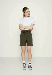 Mulher a usar uma t-shirt branca, calções olivais de cintura alta com um laço, meias brancas e ténis pretos estilo high-top, de pé com os braços cruzados.