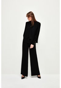 Blusa nera a maniche lunghe con fiocco, abbinata a pantaloni neri larghi. Presenta dettagli con bottoni dorati sui polsini.