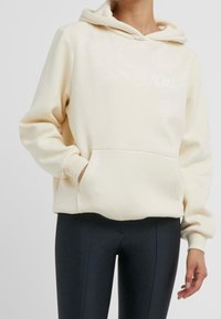 Felpa crema con tasca a marsupio, polsini a coste e design semplice. Presenta testo bianco sul davanti, abbinato a leggings neri.