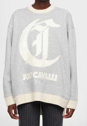 Person iført en lys grå oversize sweatshirt med cremefarvede ribkantede ærmer og kant, med stort "Just Cavalli Milano" logo foran.