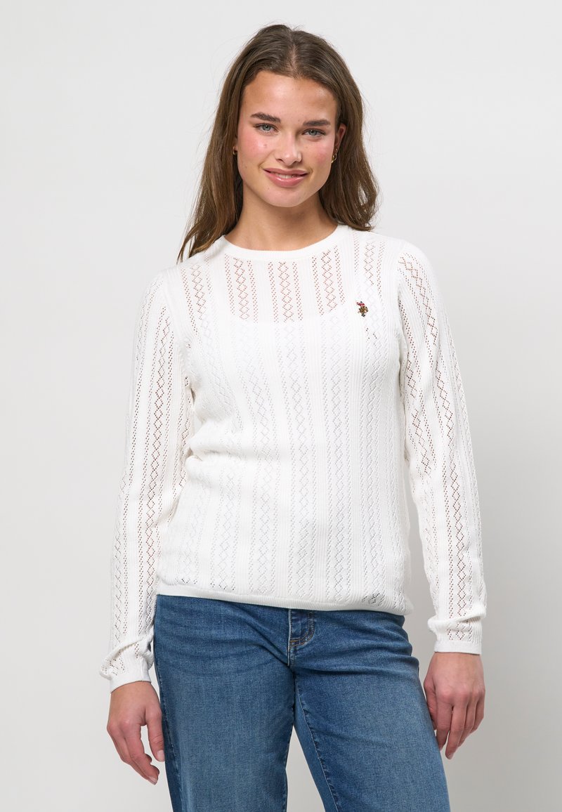 Jeune femme aux longs cheveux bruns portant un haut blanc à manches longues en dentelle et un jean bleu, souriante devant un fond clair uni.