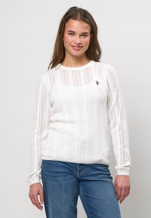 Jauna moteris su ilgais rudais plaukais, dėvinti baltą nėriniuotą ilgomis rankovėmis viršutinę dalį ir mėlynas džinsines kelnes, šypsosi priešais paprastą šviesią foną.