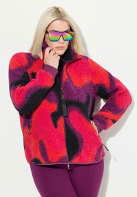 Veste en polaire présentant un motif abstrait audacieux en rouge, violet et noir. Col montant, fermeture éclair à l'avant et poches latérales pour plus de fonctionnalité.