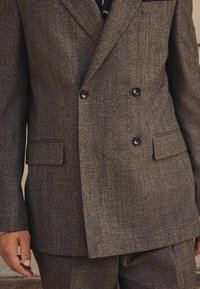 Hnědý dvojřadý blazer s houndstooth vzorem vyrobený z texturované tkaniny, s dvěma předními kapsami a černými knoflíky. Kalhoty viditelné pod ním.