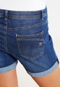 Denimshorts i mörkblått med upprullade fållar. Har två bakfickor och en broderad logodetalj. Texturen verkar slät och avslappnad.