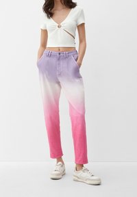 Bílý crop top s žebrováním a detailem na twist, spárovaný s joggingovými kalhotami v ombre přechodu z fialové do růžové, které mají kapsy a uvolněný střih, doplněný béžovými teniskami.
