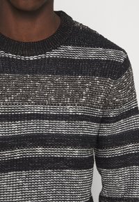 Stickad tröja med horisontella ränder i svart, grått och vitt. Rund hals med texturerad tyg och synliga sömnadsdetaljer.