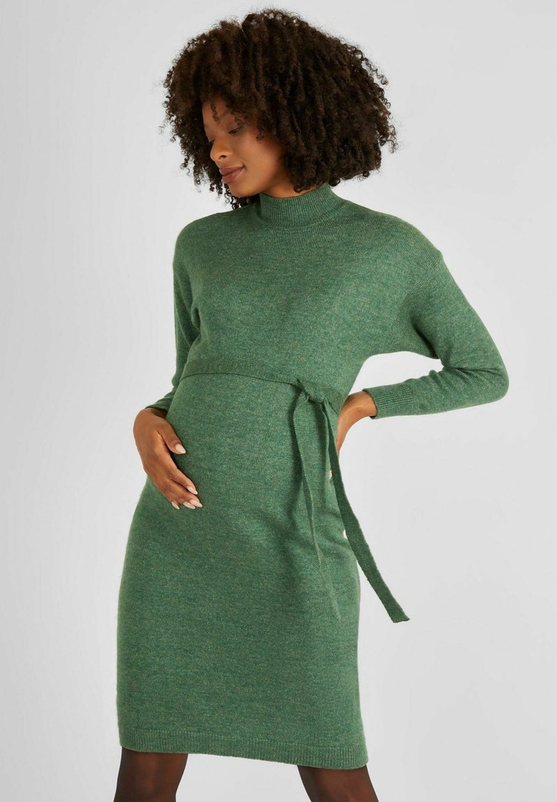 Abito lavorato a maglia verde con scollo alto, maniche lunghe e cintura con nodo in vita. La texture è morbida e leggermente strutturata, con una silhouette aderente.