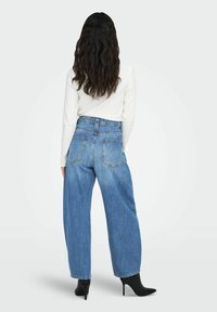 Traperice s visokim strukom, širokih nogavica, u plavoj boji, s vidljivim džepovima i lagano izblijedjelim izgledom, u kombinaciji s crnim čizmama na petu i dugom, valovitom kosom.