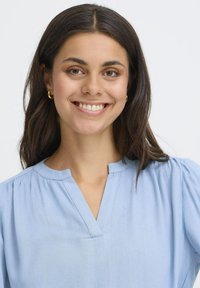 Jeune femme aux cheveux bruns, portant des boucles d'oreilles en or et une blouse bleu clair, souriant devant un fond blanc uni.
