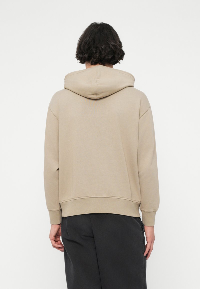 Beige hooded sweatshirt with a loose fit, ribbed cuffs, and hem. Soft texture, no visible logos or patterns. Worn with dark trousers.