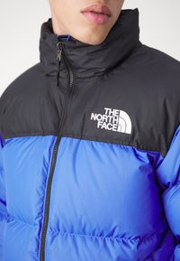 Personne portant une doudoune North Face bleue et noire, zippée jusqu'au cou, affichant le logo de la marque sur la poitrine.