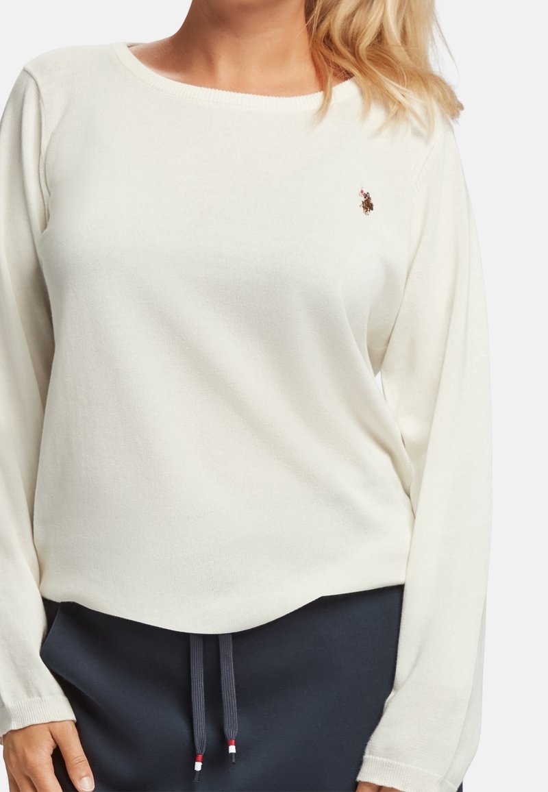 Pull en maille couleur crème avec manches longues, encolure arrondie et petit logo sur le côté gauche de la poitrine. Texture douce, coupe décontractée.