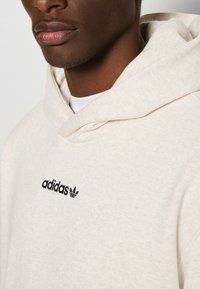 Beige Baumwoll-Hoodie mit einer vorderen Kängurutasche, einer Kapuze mit Kordelzug und einem schwarzen, gestickten Adidas-Logo auf der Brust.