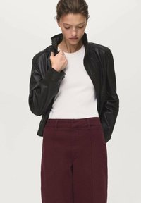 Giacca di pelle nera con colletto a zip, indossata sopra una camicia bianca, abbinata a pantaloni color amaranto. Texture liscia, design aderente, nessun motivo visibile.