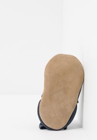 Pantof de material albastru cu talpă din suede bej, vârf rotunjit și design minimalist, sprijinit de un perete alb.